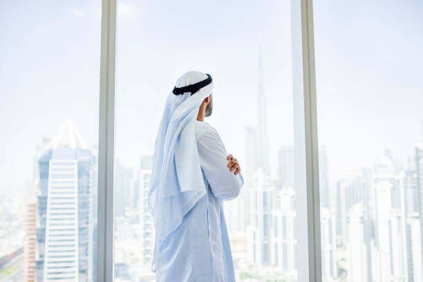 A man in traditional attire looks out a wide window over a cityscape, reflecting on Ajman Free Zone license costs.