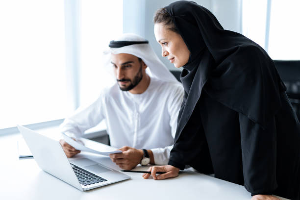 Two professionals in traditional attire review documents at a laptop, discussing Ajman Free Zone license costs.